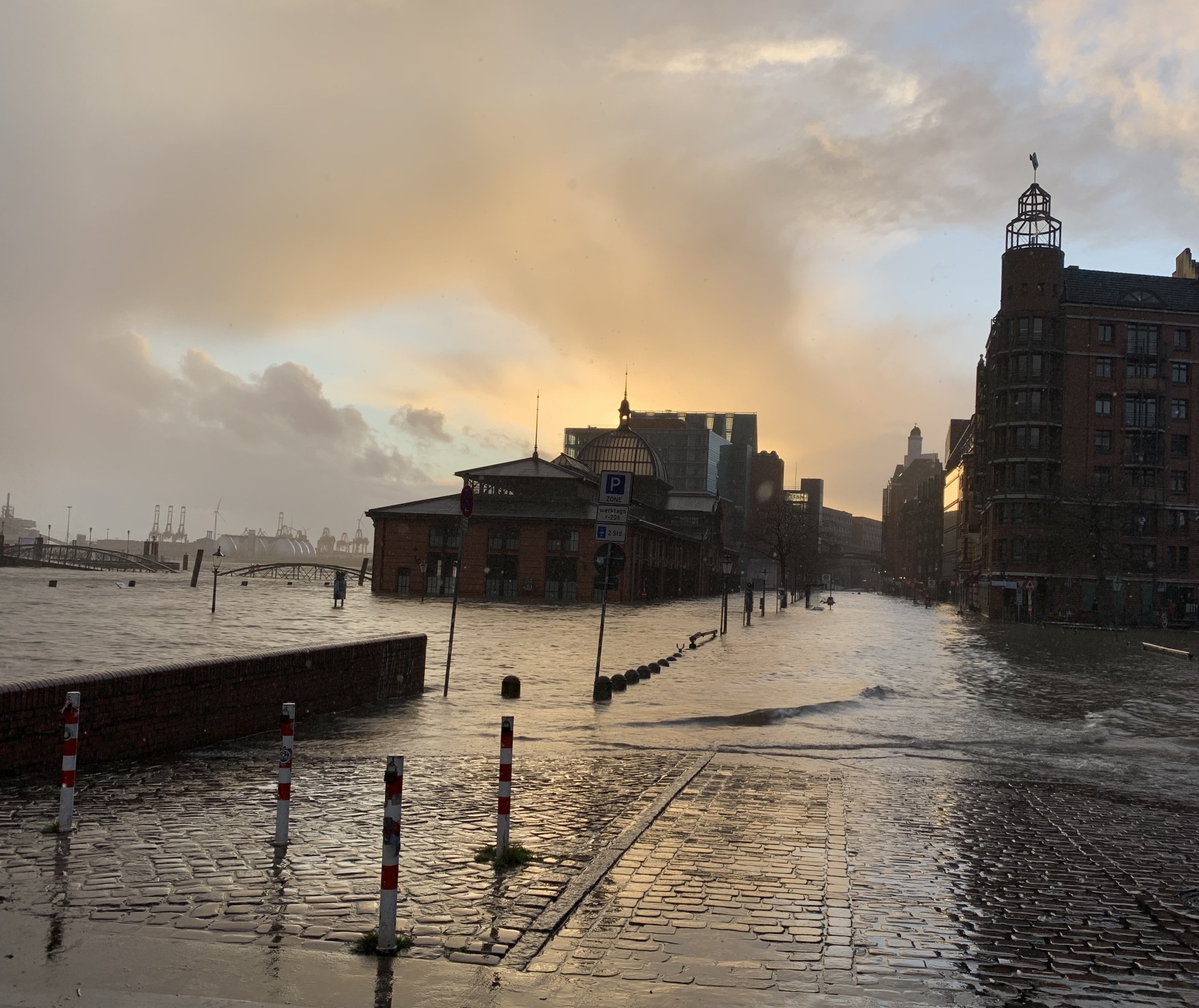 Öffentliche Straße in Hamburg, Deutschland, mit Wasser bedeckt, Masten, Schilder, Gebäude mit Fenstern, eine Brücke und ein bewölktes Himmel.