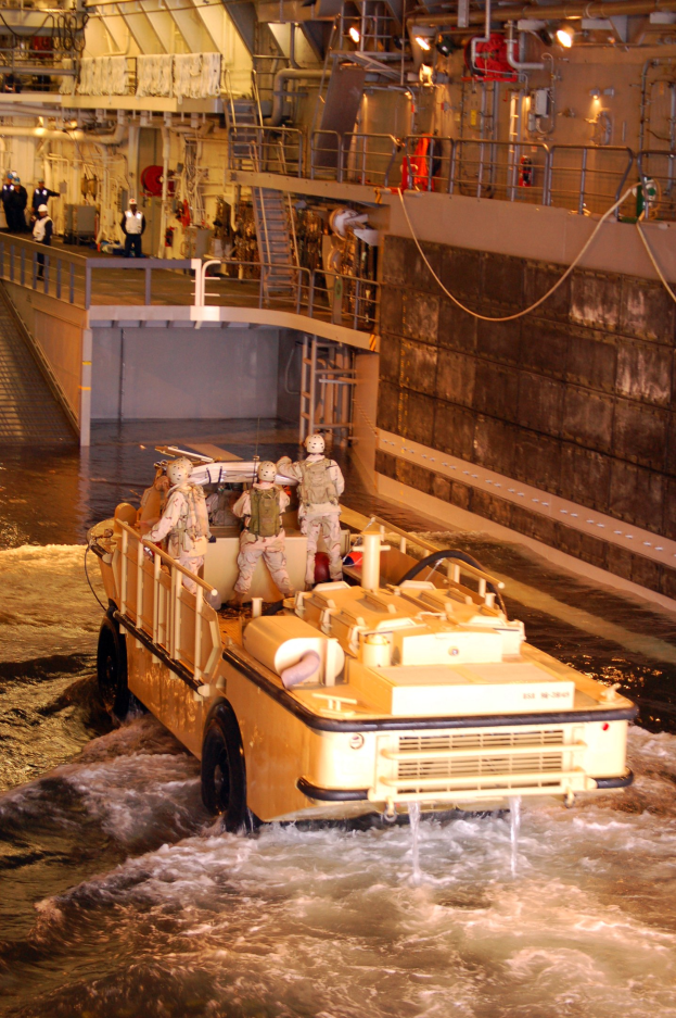 Amphibisches Fahrzeug mit Helmträgern durch Wasser neben einer Wand, mit Menschen, Geländern, Lampen und Rohren im Hintergrund.
