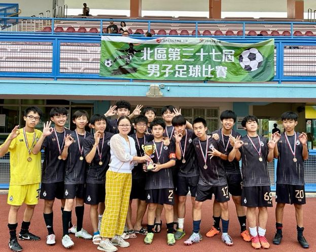 Group of young men in soccer uniforms standing on a field holding a trophy and wearing medals, with a banner for the "Yokohama U-16 Boys Soccer Team" in the background and spectators seated nearby.