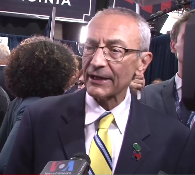 Senator Chuck Schumer in Anzug und Krawatte spricht zu Reportern bei einer Presseveranstaltung, mit Teilnehmern in Mänteln und ID-Karten im Hintergrund, während eine Tafel im Hintergrund den Text 'Sen. Schumer sagt, er sei 'kein Demokrat' anzeigt.