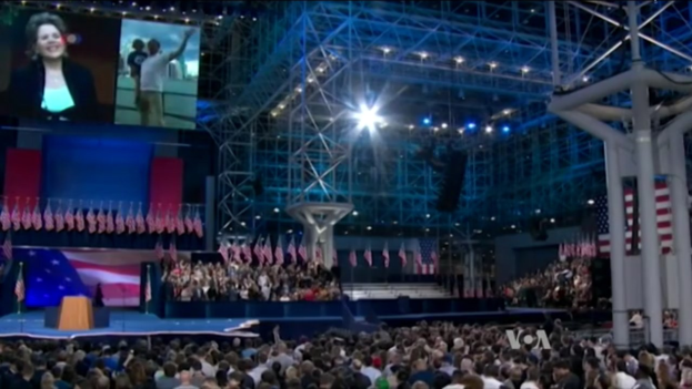Eine große Menschenmenge sitzt vor einer Bühne mit einem Podium, Mikrofon, Flaggen, einem Bildschirm und Beleuchtungsausrüstung und wartet auf eine Präsidentenrede.