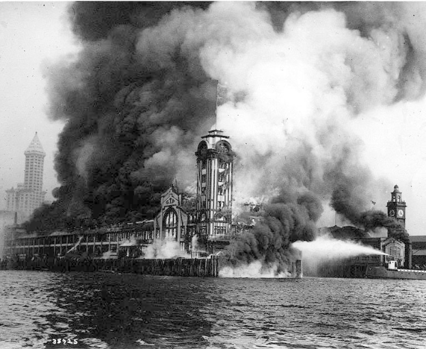 Schwarz-weiß-Foto eines großen brennenden Gebäudes mit Rauchwolken, einem Gewässer im Vordergrund und anderen Gebäuden und Himmel im Hintergrund.
