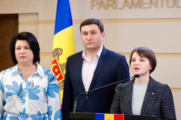 Zwei Frauen und ein Mann stehen an einem Rednerpult vor einer ukrainischen Flagge, wobei der Mann einen schwarzen Blazer, ein weißes Hemd und schwarze Hosen trägt und die Frau rechts ein graues Kleid trägt, vor einer cremefarbenen Wand mit Text und einer braunen Tür links.