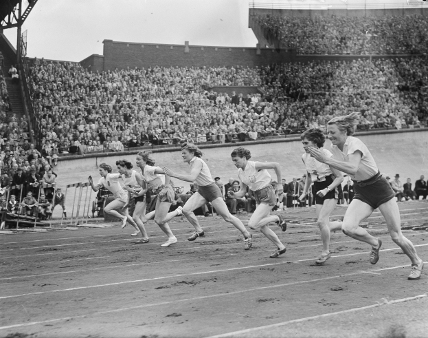 Eine Gruppe von Frauen, die auf einer Laufbahn in einem Stadion laufen, mit einer Zuschauermenge im Hintergrund, alles in Schwarz-Weiß dargestellt.