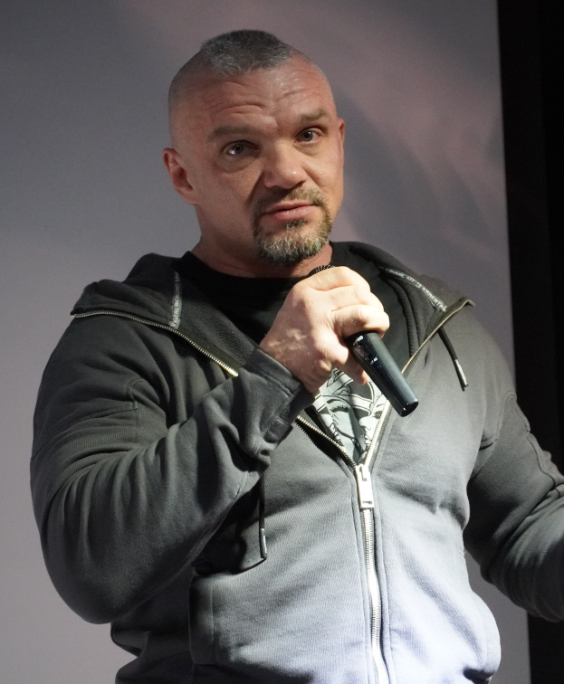 Ein Mann in einer schwarzen Kapuzenjacke und einem schwarzen Anzug mit weißem Hemd, der ein Mikrofon hält, steht selbstbewusst vor einer Wand und sieht aus wie ein Komiker mit zurückgekämmtem Haar.