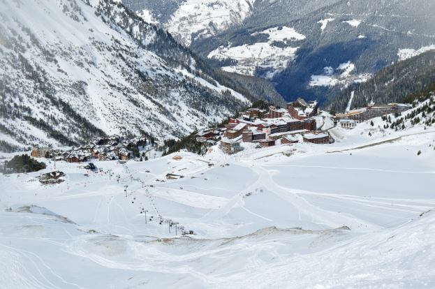 Ein Skigebiet in schneebedeckten Bergen mit Bäumen und Gebäuden im Vordergrund und einem klaren blauen Himmel im Hintergrund.