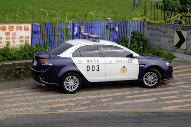 Polizeiauto am Straßenrand mit umgebender Vegetation, einem Zaun, einem Schild, einer Wand, einem Strommast, einer Satellitenschüssel und Bäumen.
