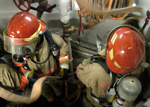 Feuerwehrleute in Helmen und Schutzausrüstung arbeiten an einem Feuerhydranten mit Rohren, Ventilen und anderen Gegenständen im Hintergrund, mit Text am unteren Bildrand.