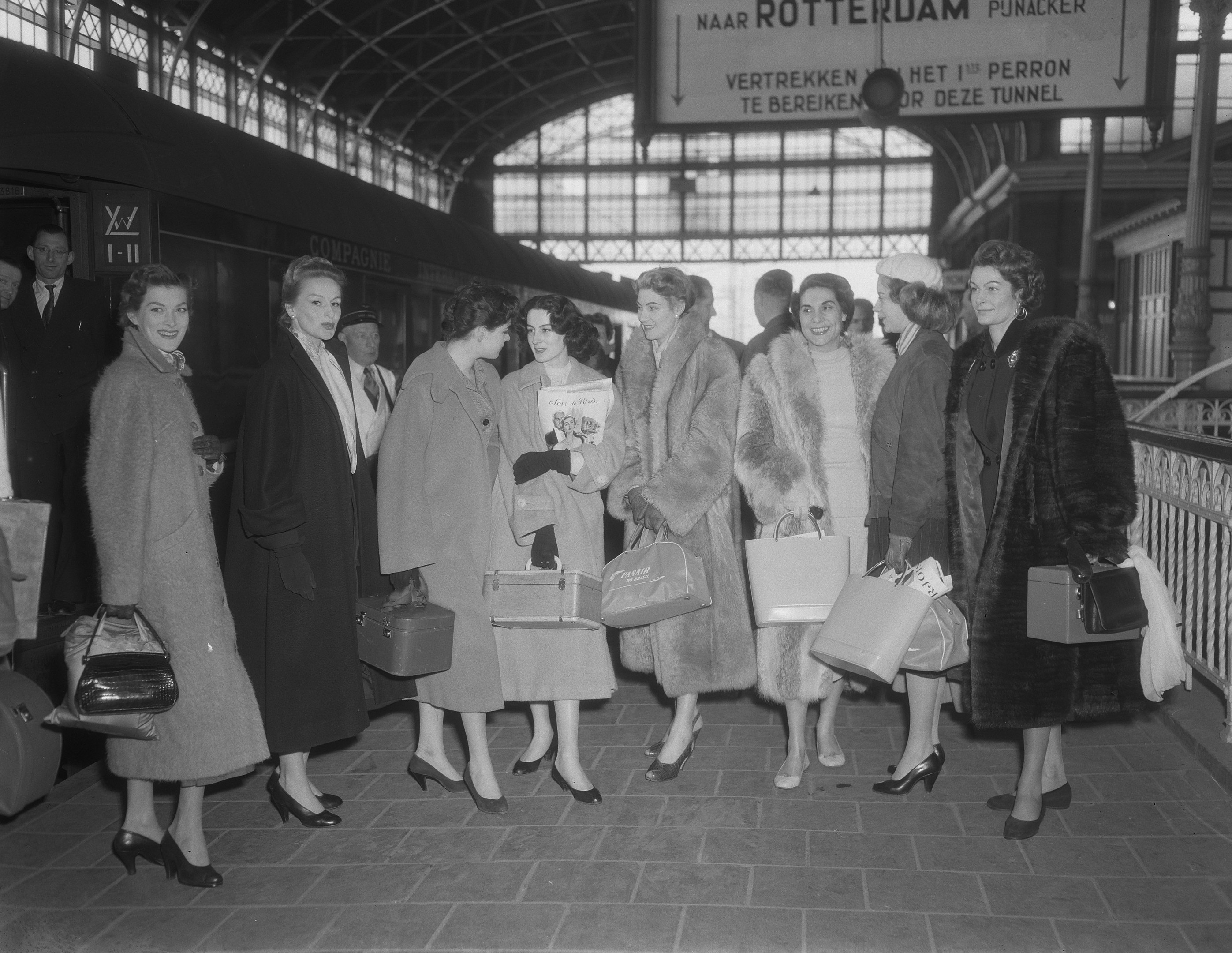 Black and white image of a group of women standing at a train station holding bags, with a train on the left and railings and a board on the right.