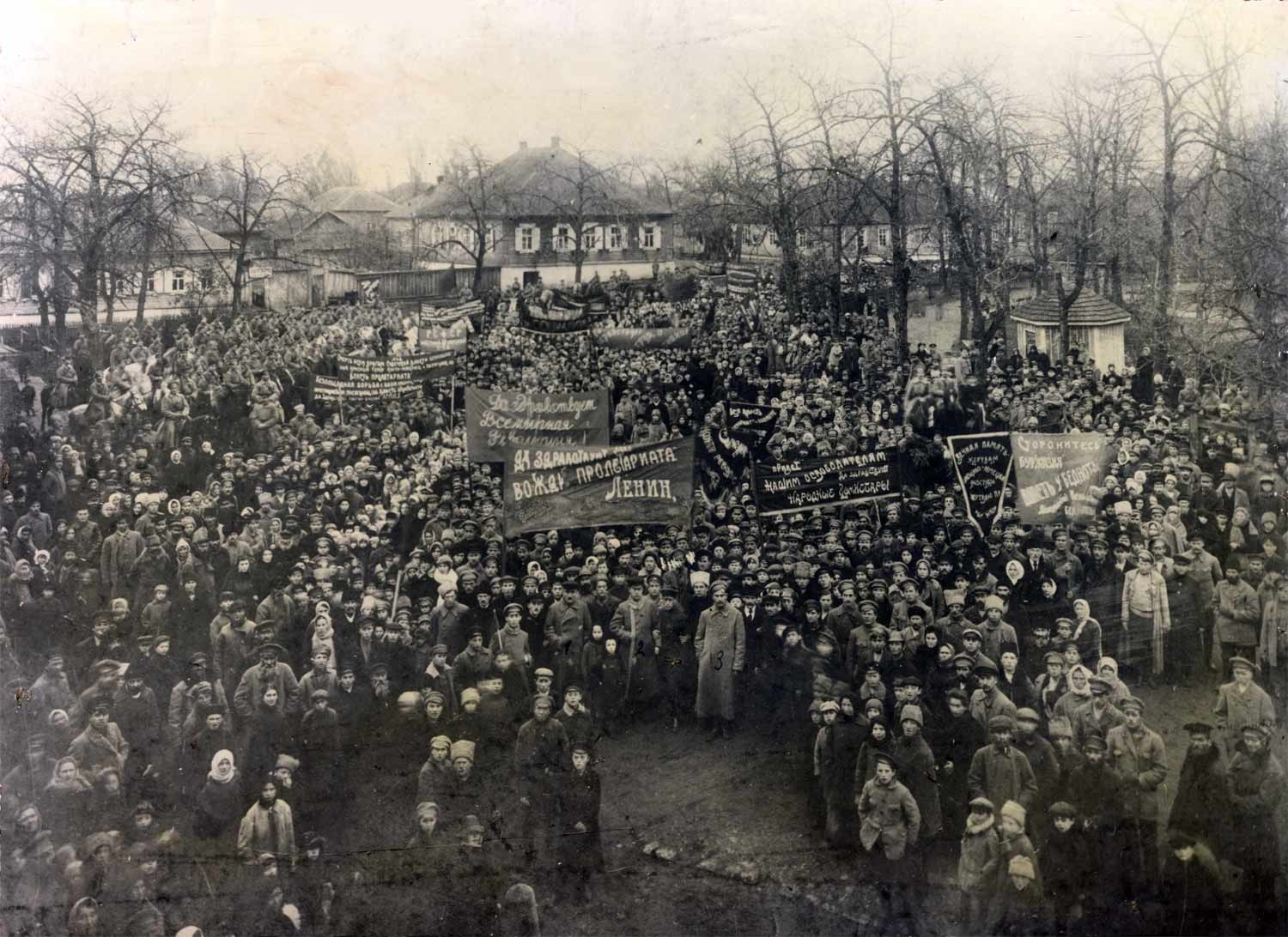 Black and white image of a large crowd holding banners in front of a building, with trees and windows in the background, suggesting a protest against the Soviet Union.