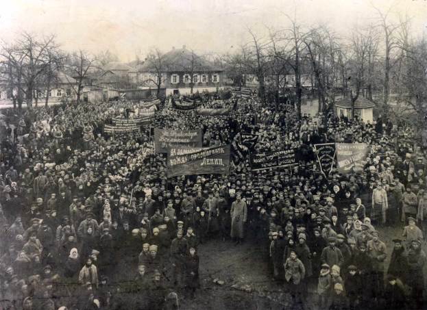 Black and white image of a large crowd holding banners in front of a building, with trees and windows in the background, suggesting a protest against the Soviet Union.