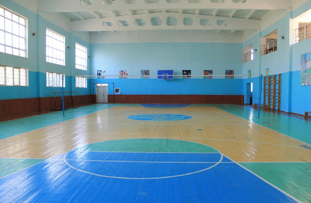 Große Turnhalle mit einem blauen und weißen Basketballfeld, Netz, Stangen, einer Tür, Fenstern, Plakaten an den Wänden, einem Geländer und einem Dach.