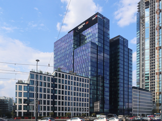 Außenansicht des Deutschen Bank-Hauptsitzes in Frankfurt, Deutschland, mit einem mehrstöckigen Gebäude mit zahlreichen Fenstern, umgeben von städtischer Infrastruktur, einschließlich Straßenlaternen, Fahrzeugen, Grünflächen und einem bewölkten Himmel.