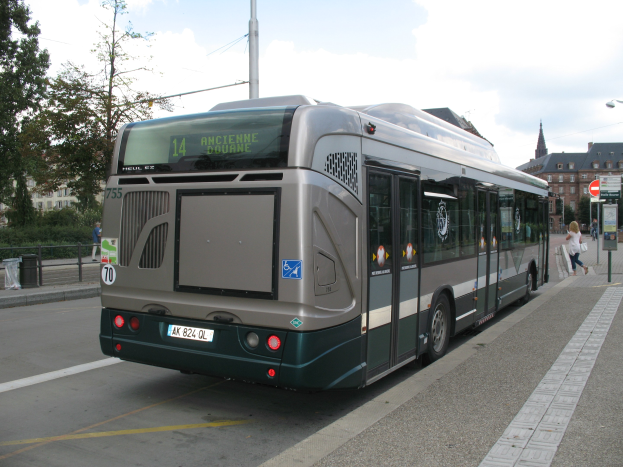Eine Stadtbus fährt auf einer von hohen Gebäuden gesäumten Straße, mit Fußgängern auf einem Gehweg und Schildern auf Pfosten auf der rechten Seite und Bäumen, Gebäuden und Himmel im Hintergrund.