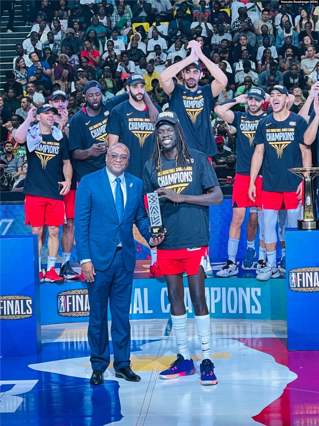 A group of men holding trophies on a basketball court, with spectators seated in the background and two trophies on podiums to the right.