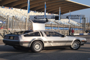 Ein silberner DeLorean DMC-12 fährt auf der Straße umgeben von Menschen, Flaggen, Schildern, Geländern, Stühlen und einem Schuppen, mit Bäumen und einem klaren blauen Himmel im Hintergrund.