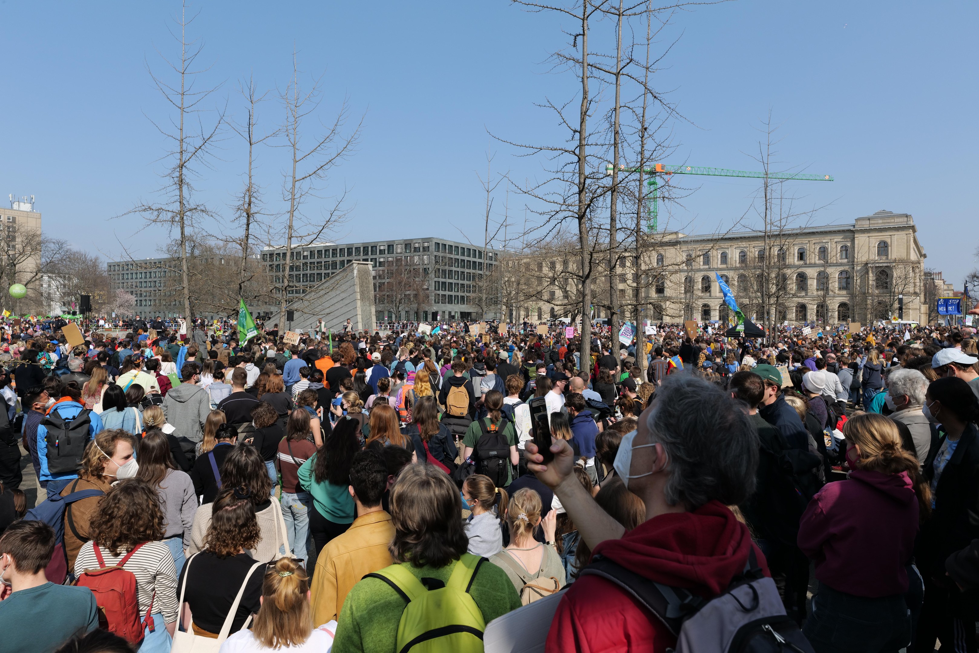 A large crowd of people stands in front of a building with windows, trees, and a clear sky, many holding placards and wearing bags, indicating a climate change protest.