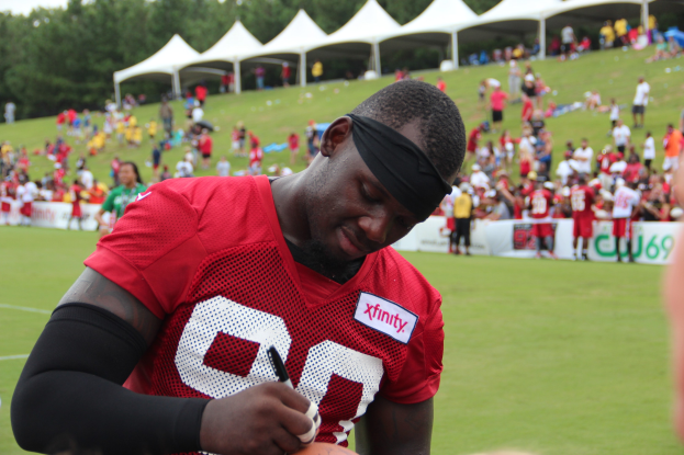 A man in a red shirt signing a football on a field with a crowd, banners, tents, trees, and a clear blue sky.