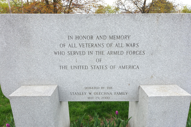 Denkmalstein mit einer Hommage an Veteranen der US-Streitkraefte, umgeben von Pflanzen, Blueten, Gras, Baeumen und einem bewölkten Himmel.
