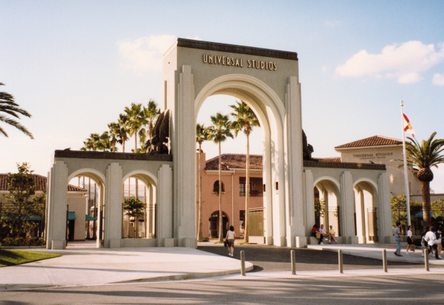 Eingang zu Universal Studios Hollywood mit einem Bogen mit Text, einer Statue, Menschen, Bäumen, Gebäuden, einem Fahnenmast mit einer Fahne.