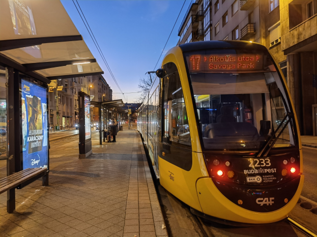 Gelber Straßenbahnwagen auf einer nächtlichen Stadtstraße mit einer Bank und einem Bushaltestellenschild auf der linken Seite, Gebäuden, Straßenlaternen, Bäumen und dem Nachthimmel im Hintergrund.