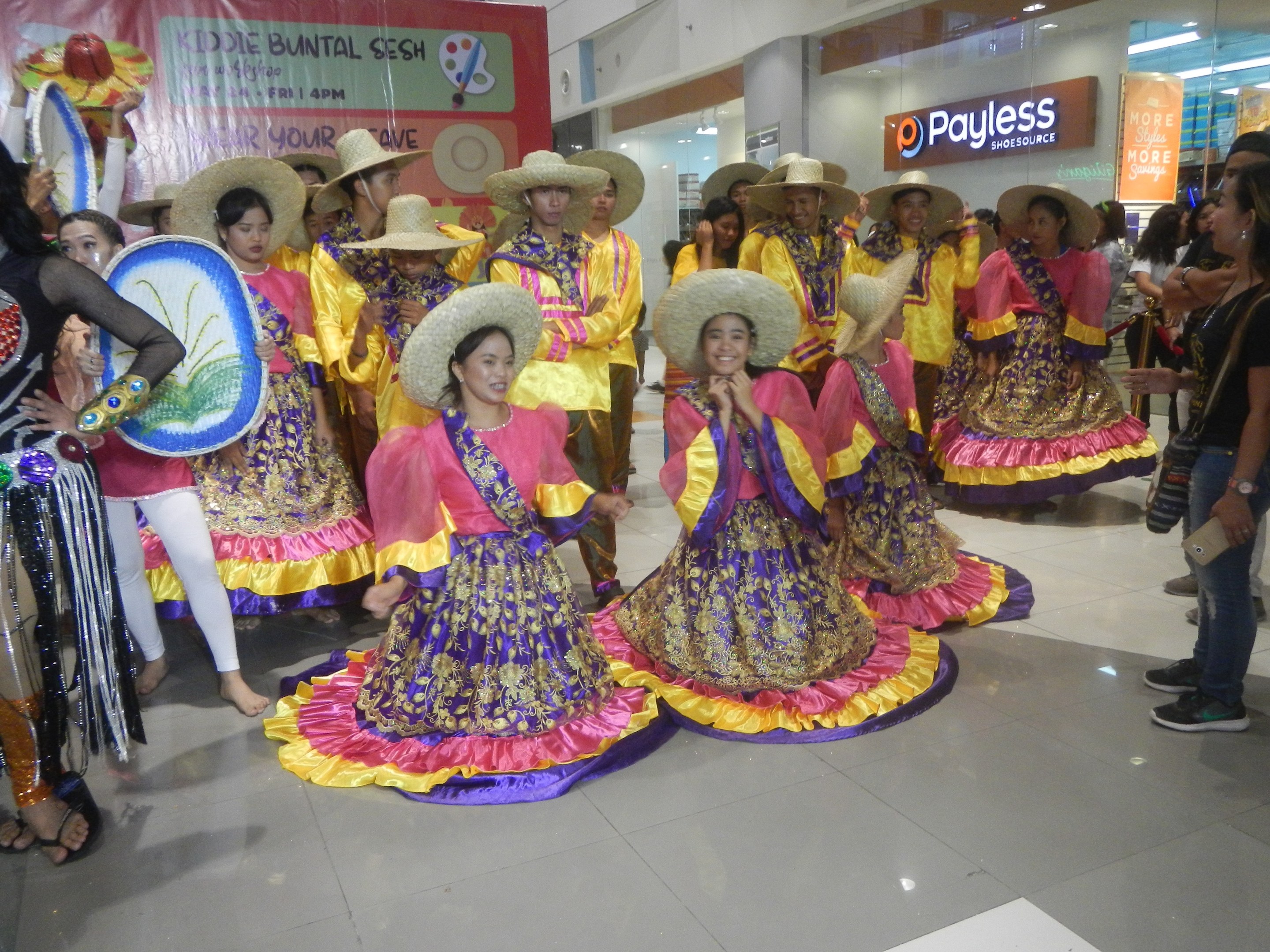 Eine Gruppe von Menschen in bunten Kleidern und Hüten, die in einem Einkaufszentrum während eines festlichen Ereignisses tanzen, mit anderen, die in der Nähe stehen und Musikinstrumente sowie ein Schild im Hintergrund halten.