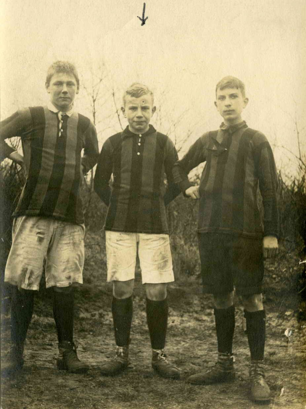 Drei junge Männer in Fußballtrikots stehen auf einem Feld mit Bäumen und einem klaren Himmel im Hintergrund.