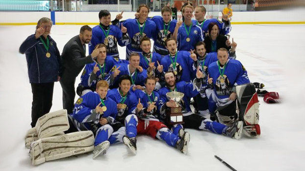 Eine Gruppe von Männern feiert einen Meisterschaftssieg auf einem Eisstadion, trägt Medaillen und hält einen Pokal, mit Zuschauern und Glaswänden im Hintergrund.