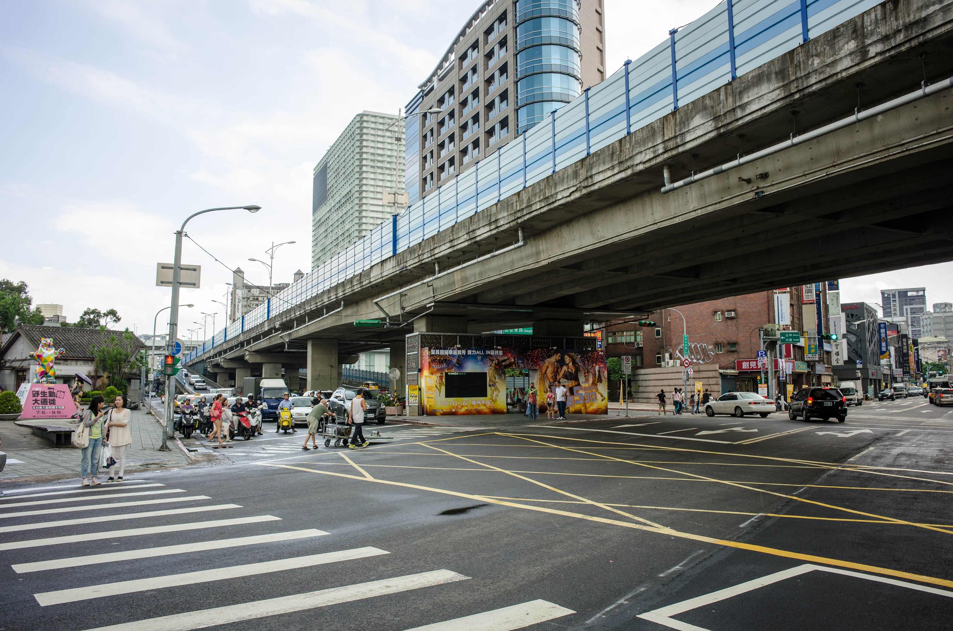 A bustling city street with a pedestrian bridge overhead, vehicles on the road, pedestrians on the sidewalk, buildings with windows in the background, lined with light poles under a cloudy sky.