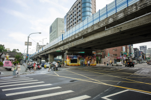 A bustling city street with a pedestrian bridge overhead, vehicles on the road, pedestrians on the sidewalk, buildings with windows in the background, lined with light poles under a cloudy sky.