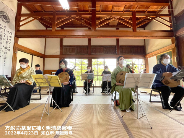 Eine Gruppe von Menschen mit Masken spielt Musikinstrumente drinnen, mit Bücherständern, einem Banner, Fenstern, Deckenlampen und Text, der ein japanisches Musikfestival anzeigt.