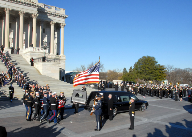 A funeral procession with people in caps and flags marches past the U.S. Capitol Building, flanked by vehicles, steps, railings, light poles, and trees under a sky.