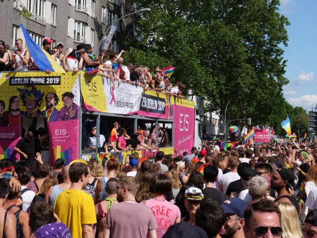 Eine große Menge marschiert während des Berlin Pride 2019 die Straße entlang, viele tragen Mützen und Schutzbrillen, einige halten Fahnen, mit Bannern, Laternenpfählen, Bäumen und Gebäuden im Hintergrund unter einem bewölkten Himmel.