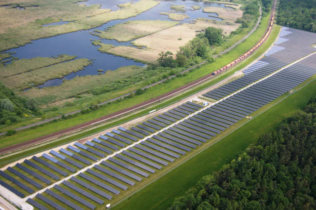 Eine Luftaufnahme eines Solarparks mit zahlreichen Solarmodulen auf einem Feld, umgeben von Bäumen, Gras, Wasser und einer Bahnlinie in der Nähe.