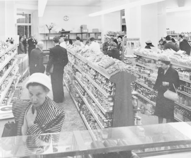 Schwarzes und weißes Foto von Menschen beim Einkaufen in einem Supermarkt mit Lebensmittelregalen, einige halten Taschen, eine Wanduhr und Deckenbeleuchtung.