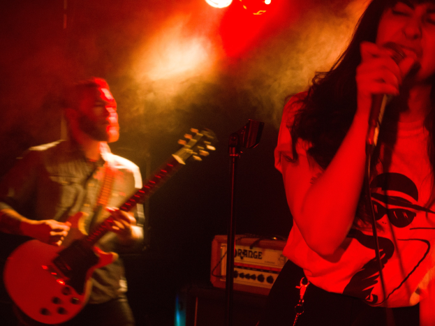 Eine Frau singt in ein Mikrofon auf der Bühne, während ein Mann im Hintergrund eine akustische Gitarre spielt, mit Deckenlampen, die die Szenerie erhellen.