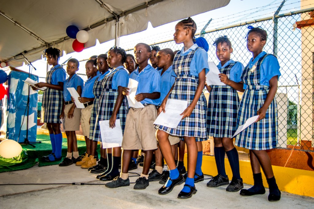 School children in uniforms stand on a path holding papers, with one girl speaking into a microphone on a stand, while a fencing wall with a shed topped by balloons is visible behind them.