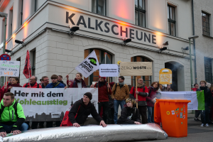 Eine Gruppe von Menschen hält Schilder und Plakate vor einem Gebäude, während zwei Personen im Vordergrund sitzen und ein Müllcontainer rechts zu sehen ist, während einer Demonstration in Deutschland, mit Gebäuden, Fenstern, Lichtern und Schildern im Hintergrund.