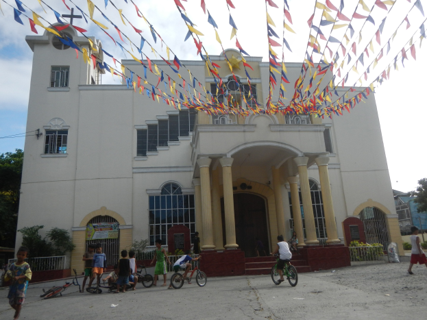 Gruppe von Menschen, die vor einer weißen Kirche mit architektonischen Details wie Fenstern, Säulen, Bögen, Türen und einem Kreuzsymbol Fahrrad fahren, mit Pflanzen, einem Zaun, einer Fahne, Bäumen und einem bewölkten Himmel im Hintergrund und bunten Fahnen, die über einem Seil gebunden sind.