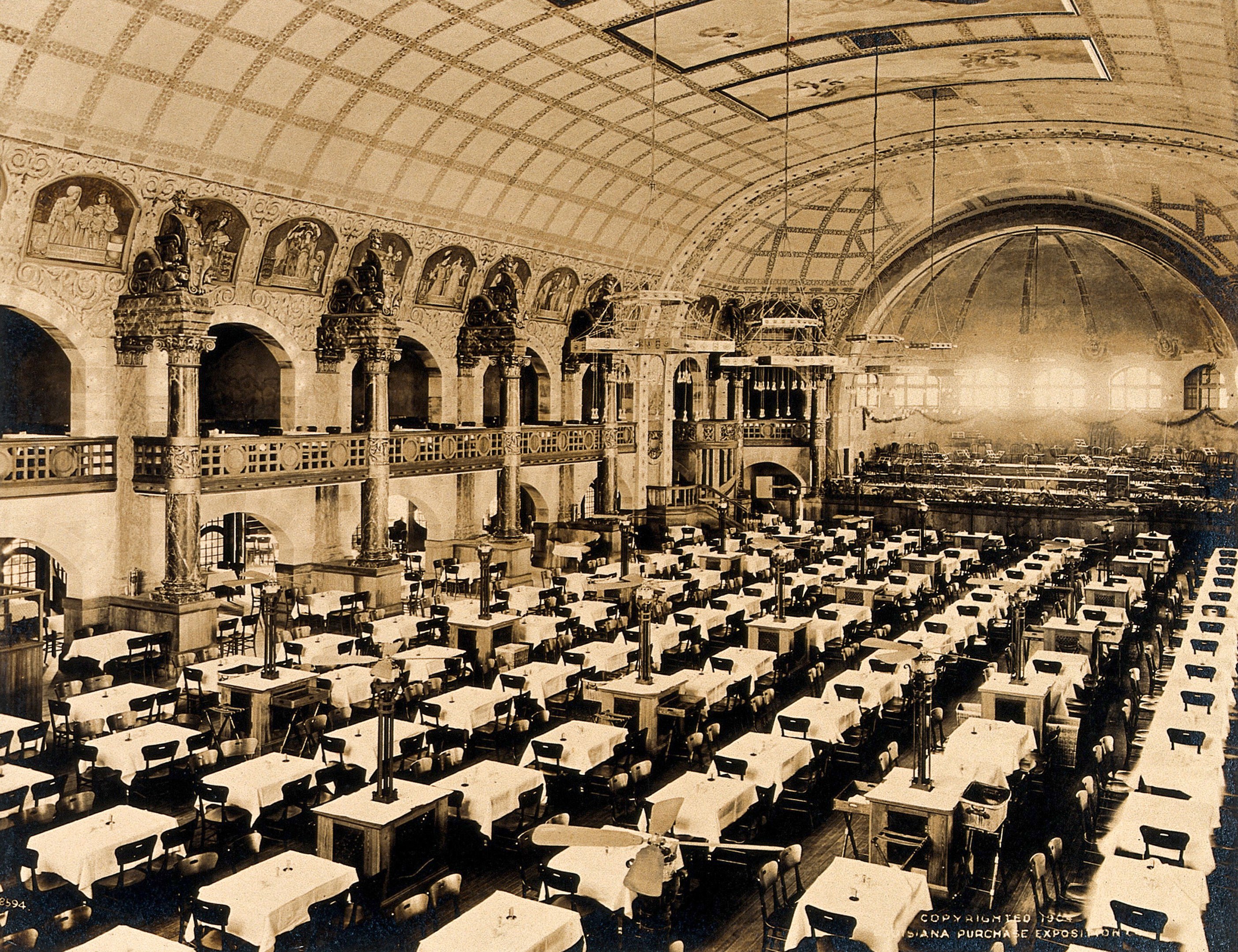 A vintage photograph of a large room with numerous tables and chairs, supported by pillars, featuring railings, wall sculptures, and ceiling lights.