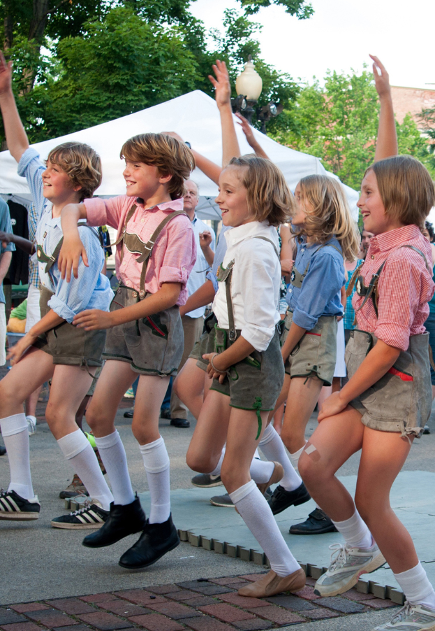 Eine Gruppe von Kindern in traditioneller bayerischer Tracht tanzt auf einer Straße, umgeben von einer Menge, Zelten, Laternenmästen, Bäumen, Gebäuden und einem klaren blauen Himmel, wobei einige Kinder Shorts und Schuhe tragen.