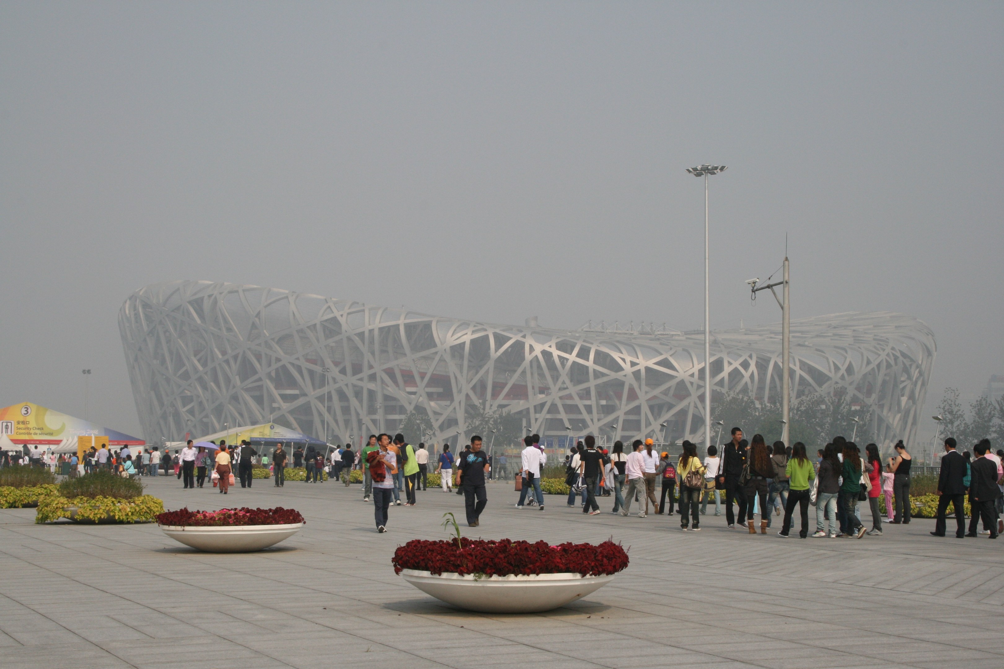 Olympiastadion in Peking, China mit Menschen, die herumlaufen, Pflanzen mit Blumen im Vordergrund, Laternenmasten, Bäume und einen klaren blauen Himmel im Hintergrund.