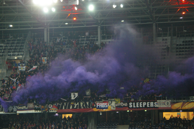 Große Menschenmenge in einem Stadion mit lila Rauch, Bannern, Stühlen, Treppen, Geländern und Deckenbeleuchtung.