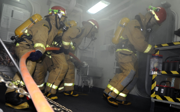 Feuerwehrleute in Schutzausrüstung mit Helmen und Sauerstoffflaschen arbeiten an einem Feuerlöschwagen, mit einem Gestell voller Gegenstände und Hintergrundausrüstung im Hintergrund.