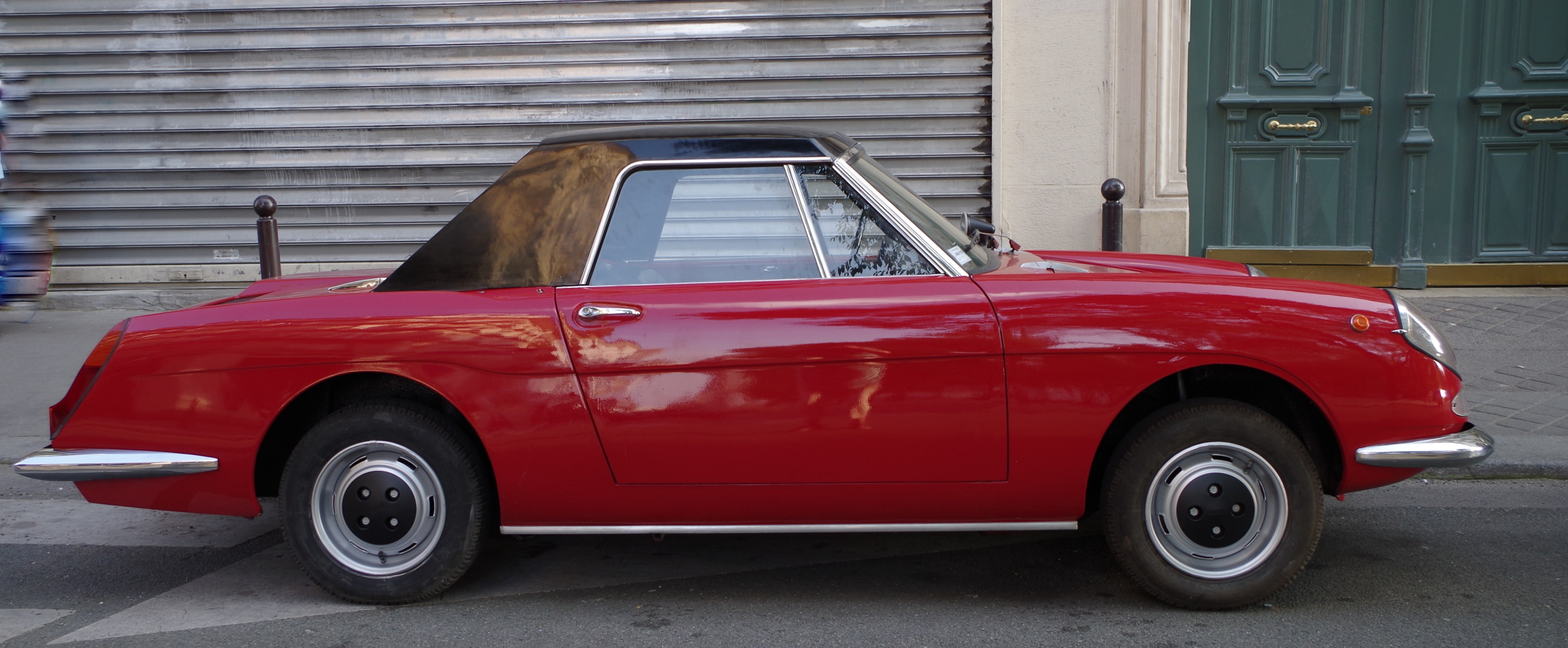 Rotes Auto auf der Straßenseite geparkt mit einer Person, die auf dem Gehweg zu seiner Linken geht, Verschluss und grüne Tür im Hintergrund.