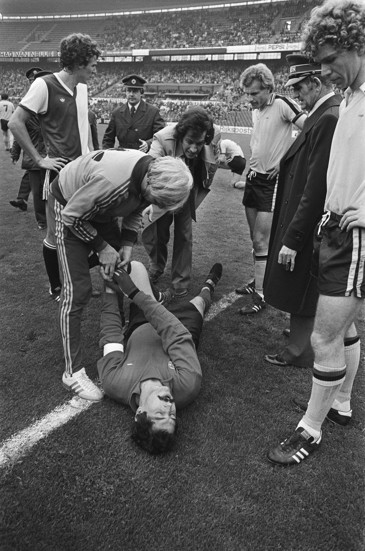 Schwarzes und weißes Bild eines Fußballspielers, der auf dem Boden liegt und von einer Gruppe Männer umgeben ist, mit einer Menge im Stadionhintergrund; der Spieler scheint verletzt zu sein.
