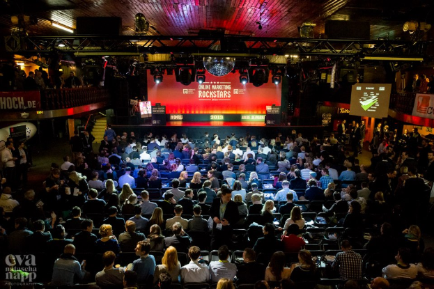 Eine große Menschenmenge sitzt auf Stühlen vor einer Bühne mit Bildschirmen und Bannern, die von Scheinwerfern beleuchtet wird und von Geländern umgeben ist, und wartet gespannt auf den Beginn eines Online-Marketing-Rockstars-Events, mit einem Wasserzeichen in der linken unteren Ecke.