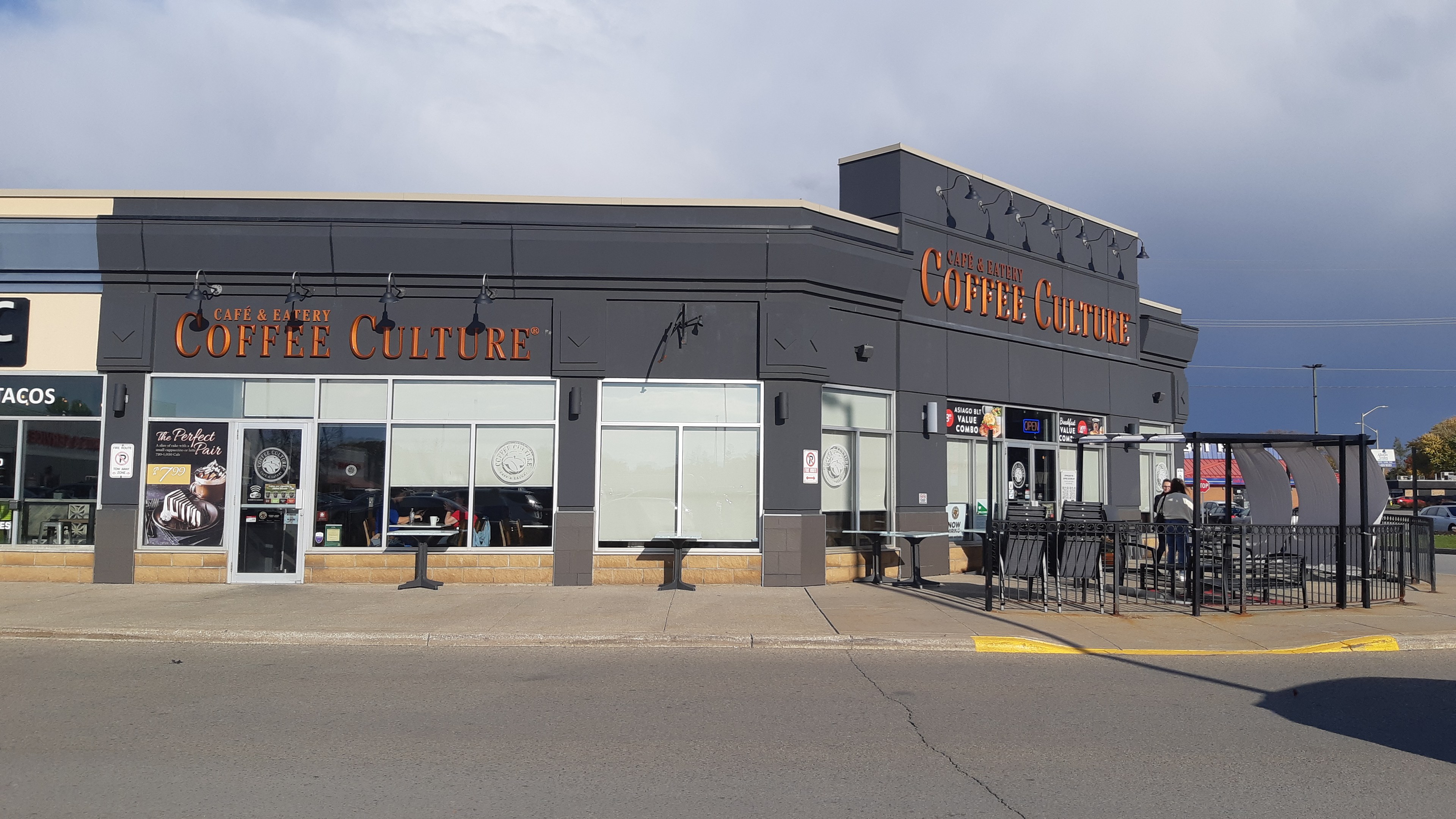 Ein Kaffeeladen an einer Straßenecke mit einer Straße im Vordergrund, einem Gebäude im Hintergrund, Fahrzeugen, Bäumen und einem blauen und weißen Himmel.