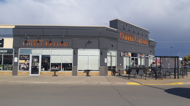 Ein Kaffeeladen an einer Straßenecke mit einer Straße im Vordergrund, einem Gebäude im Hintergrund, Fahrzeugen, Bäumen und einem blauen und weißen Himmel.