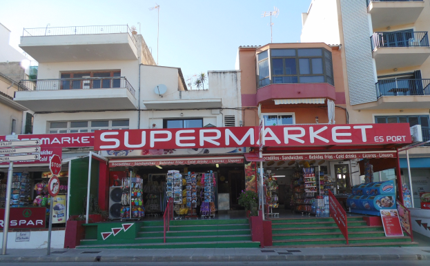 A supermarket at a street corner with buildings in the background, featuring various signs, poles, plants, and other urban elements under a visible sky.